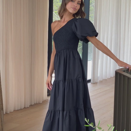 A smiling woman wearing a black one-shoulder midi dress made of crepe fabric, with a smocked bodice and a tiered skirt, standing on a light wooden floor with white curtains in the background.