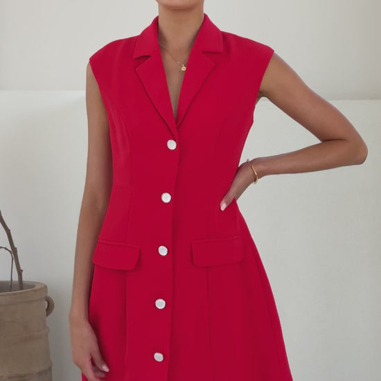 A woman wearing a vibrant red sleeveless blazer dress made of banana crepe, featuring white buttons down the front and two front flap pockets.