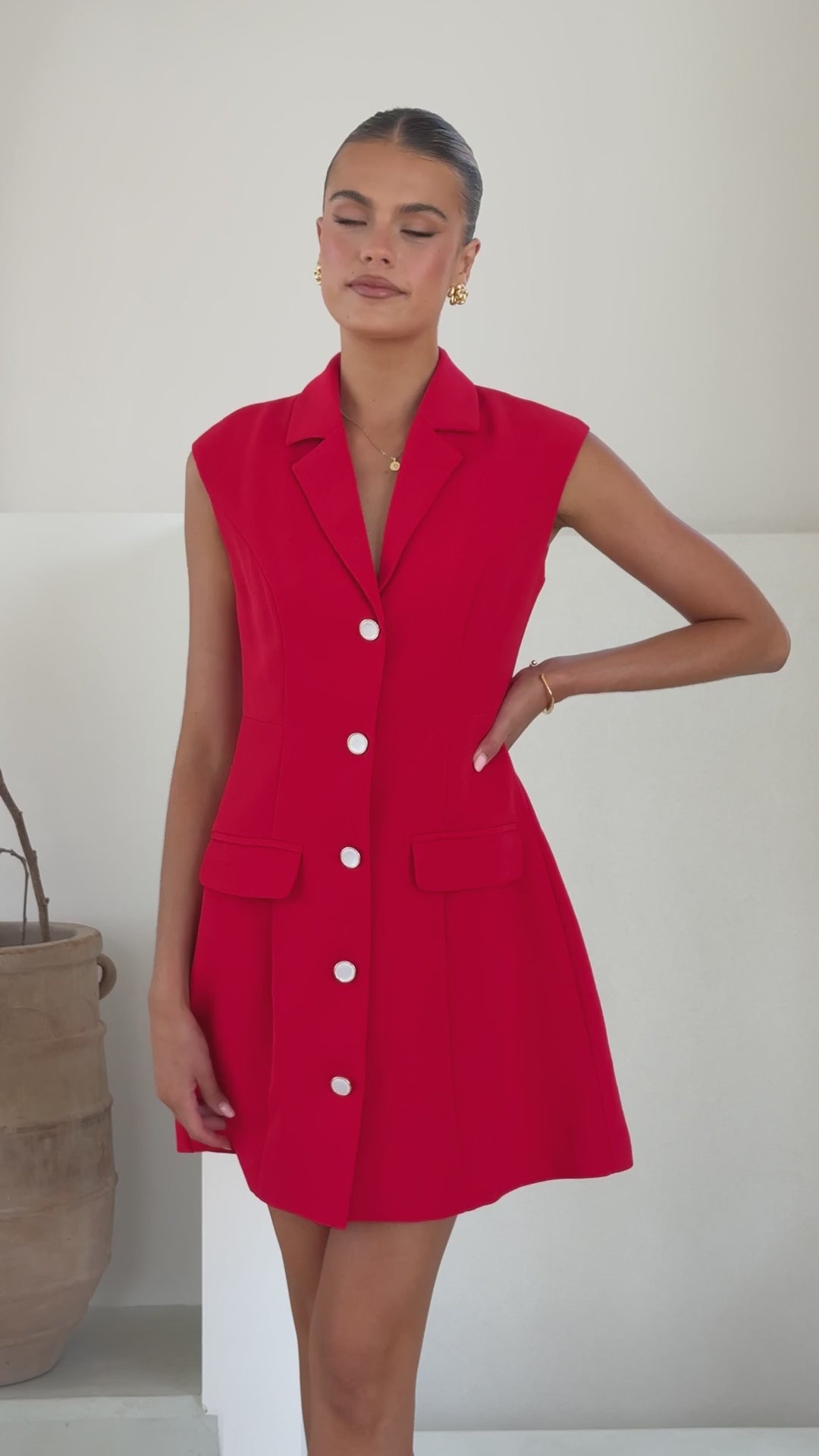 A woman wearing a vibrant red sleeveless blazer dress made of banana crepe, featuring white buttons down the front and two front flap pockets.